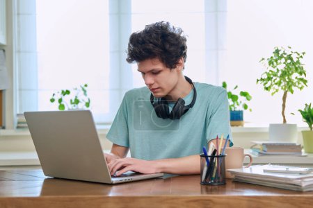 Foto de Joven estudiante universitario sentado en el escritorio en casa usando la computadora portátil. Guy 19-20 años estudiando en casa, utilizando un ordenador para la educación ocio comunicación - Imagen libre de derechos