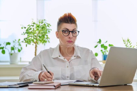 Foto de Retrato de una mujer de negocios madura que trabaja en casa en un ordenador portátil. Sonriente hembra de mediana edad mirando a la cámara sentada en el escritorio de la oficina en casa. Trabajo remoto de negocios, carrera, gestión, marketing - Imagen libre de derechos