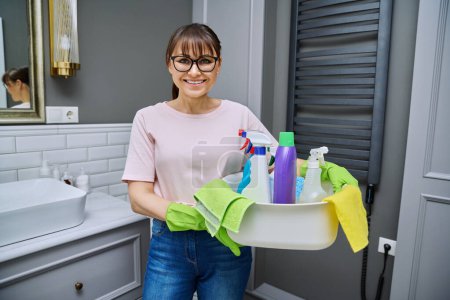 Foto de Mujer de mediana edad en guantes con lavabo de detergentes en el baño. Mujer preparándose para la limpieza rutinaria de la casa, empleada de servicio de limpieza posando en el lugar de trabajo. concepto de limpieza de las tareas domésticas - Imagen libre de derechos