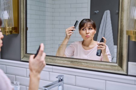Téléchargez les photos : Femme d'âge moyen avec spray réglage pour coiffer peigne de fixation des cheveux regardant dans le miroir, faire coiffure, dans la salle de bain. Beauté, âge, cosmétiques, accessoires capillaires - en image libre de droit