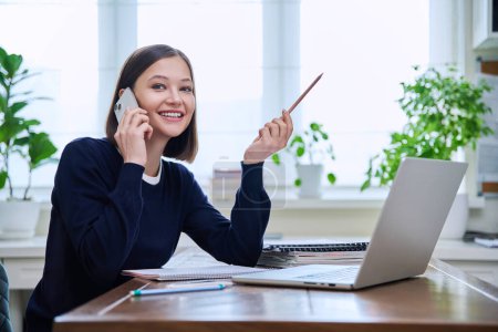 Téléchargez les photos : Joyeux sourire jeune femme assise au bureau à la maison avec ordinateur portable et parler sur smartphone. Femme travaillant, étudiant à distance, pigiste, enseignante, étudiante - en image libre de droit