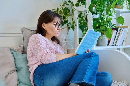 Photo for Mature woman reading paper book sitting on couch at home. Relaxation, literature hobby, fiction, lifestyle, people concept - Royalty Free Image