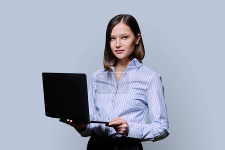 Foto de Mujer de negocios serio joven con portátil sobre fondo gris. Confiada exitosa mujer sosteniendo el ordenador en las manos mirando a la cámara. Negocios, trabajo, formación, educación, tecnologías digitales modernas - Imagen libre de derechos