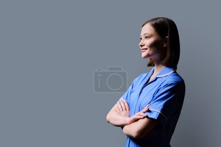 Portrait of young confident smiling female nurse with crossed arms, looking in profile on gray studio background, copy space. Medical services, health, professional assistance, medical care concept