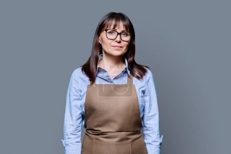 Confident serious middle-aged woman in apron on gray background. Successful mature female small business owner, service worker, entrepreneur looking at camera. Business work people startup advertising