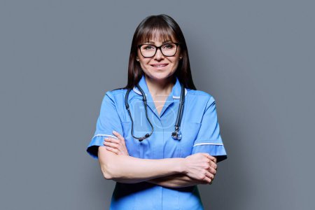 Porträt einer lächelnden Krankenschwester mittleren Alters in Blau mit verschränkten Armen und Stethoskop auf grauem Studiohintergrund. Medizinische Dienste, nhs, Gesundheit, professionelle Hilfe, medizinisches Versorgungskonzept
