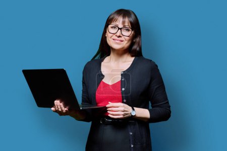 Téléchargez les photos : Femme d'âge moyen souriante d'affaires utilisant un ordinateur portable sur fond de studio bleu. Femme mature et confiante qui regarde la caméra. Entreprise, travail, emploi, style de vie, technologie, concept de personnes des années 40 - en image libre de droit