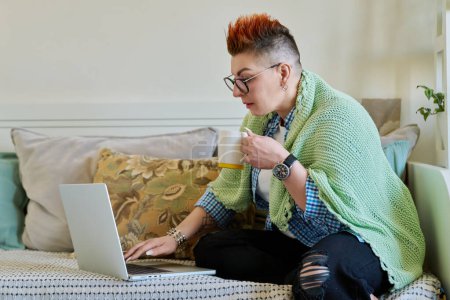 Mature woman with warm blanket holding cup of hot drink looking at laptop screen sitting on couch at home. Home coziness comfort, cold seasons, lifestyle, technology for leisure work communication