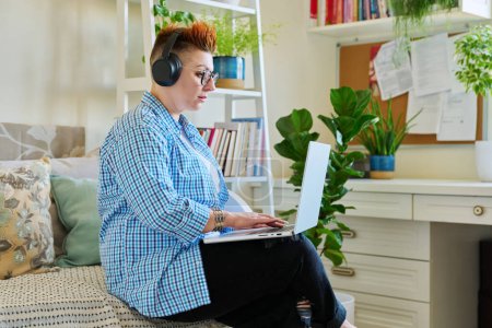 Foto de Mujer de mediana edad freelancer en auriculares con ordenador portátil sentado en el sofá en casa. 40 hembra mirando la pantalla video chat conferencia llamada reunión en línea formación mentoring tecnología para el trabajo - Imagen libre de derechos