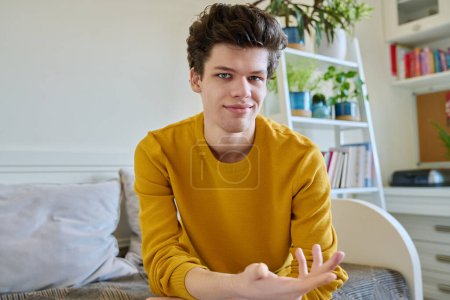 Photo for Portrait of handsome smiling guy looking at camera, web cam view, sitting on couch at home. Young man in yellow with curly hair, university college student. Lifestyle, youth 19-20 years old concept - Royalty Free Image