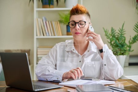 Middle-aged business woman talking on mobile phone sitting at desk with laptop computer. Serious confident mature female working with business papers. Work, workplace, 40s people