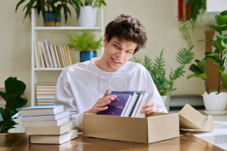 Photo for Satisfied young male customer buyer sitting at home unpacking cardboard box parcel with new books, online purchases. Delivery by mail, internet store bookstore - Royalty Free Image