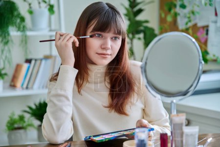 Téléchargez les photos : Jeune femme avec des produits cosmétiques sur la table, faisant maquillage regardant dans le miroir à la maison. Cosmétiques, jeunesse, mode de vie, concept beauté - en image libre de droit