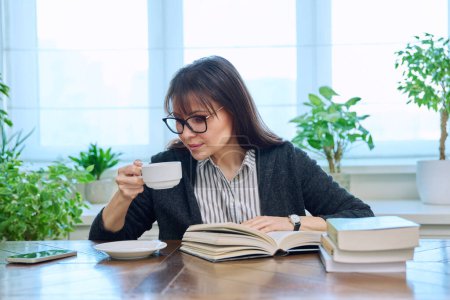 Femme d'âge moyen lisant des livres assis à la maison à une table avec une tasse de café. hobby littéraire, loisir, détente, concept de personnes