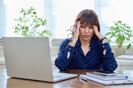 Tired sad mature woman at workplace at table with computer laptop, holding hands on temples of head. Middle age people, stress difficulty, headache health, lifestyle concept