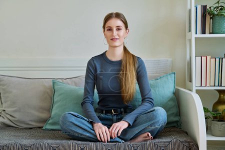 Téléchargez les photos : Portrait de heureuse jeune adolescente détendue de 17,18 ans assise en position lotus sur le canapé à la maison, souriant en regardant la caméra. Jeunesse, concept de style de vie - en image libre de droit