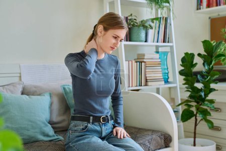 Photo for Young teenage female experiencing neck pain sitting on couch at home. Symptoms of the musculoskeletal system, neurological pathologies, acute infectious diseases - Royalty Free Image