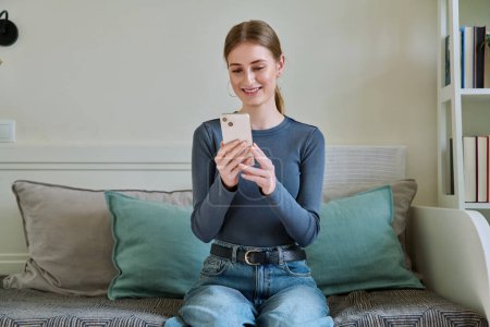 Happy smiling female teenager using smartphone, sitting on couch at home, girl 16,17, 18 years old texting reading messages. Modern digital technologies for communication, leisure, learning, shopping