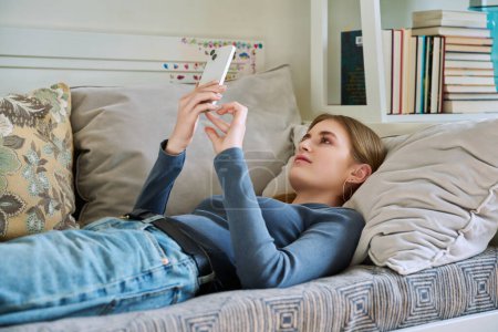 Foto de Feliz sonrisa relajada joven adolescente acostada en el sofá en casa utilizando el teléfono inteligente para la comunicación de ocio. Juventud, estilo de vida, tecnología digital en línea, gadgets, internet - Imagen libre de derechos