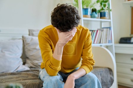 Photo for Sad upset tired young man sitting on couch at home, touching his head with hands. Health problems, headaches, troubles, difficulties in study, family relationships, mental health, stress, depression - Royalty Free Image