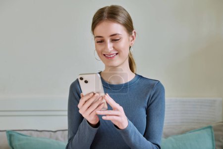 Photo for Happy smiling female teenager using smartphone, sitting on couch at home, girl 16,17, 18 years old texting reading messages. Modern digital technologies for communication, leisure, learning, shopping - Royalty Free Image