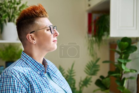 Téléchargez les photos : Portrait de profil de femme sérieuse d'âge moyen dans des lunettes avec coupe de cheveux rouge regardant la fenêtre à l'intérieur de la maison, espace de copie. Mature, mode de vie, santé, concept de vie - en image libre de droit