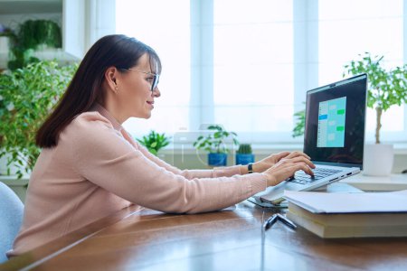 Photo for Middle-aged woman communicates online using laptop at her desk at home. Chat on computer screen, communication work leisure technology concept - Royalty Free Image