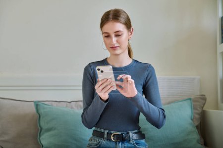 Photo for Serious calm female teenager using smartphone, sitting on couch at home, girl 16,17, 18 years old texting reading messages. Modern digital technologies for communication, leisure, learning, shopping - Royalty Free Image