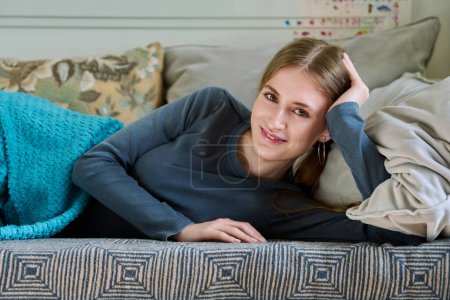 Téléchargez les photos : Belle souriante jeune adolescente calme relaxante couchée sous la couverture à la maison sur le canapé, regardant la caméra. - en image libre de droit