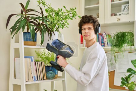 Foto de Un joven guapo que usa audífonos, aspira estanterías en casa. Limpieza, higiene, limpieza, tareas domésticas, limpieza del hogar, concepto de juventud - Imagen libre de derechos