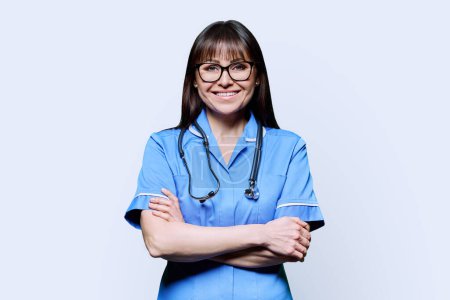 Photo for Portrait of smiling middle-aged woman nurse in blue with crossed arms with stethoscope on white studio background. Medical services, nhs, health, professional assistance, medical care concept - Royalty Free Image