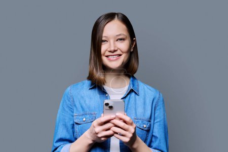 Photo for Young happy woman using smartphone in gray background. Smiling 20s female looking at camera texting. Mobile Internet apps applications technologies for work education communication shopping healthcare - Royalty Free Image