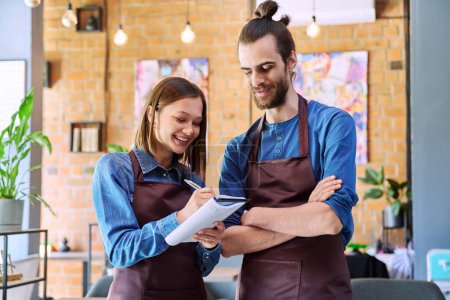 Photo for Small business team confident successful colleagues partners owners young man woman in aprons writing at working notebook at workplace in restaurant coffee shop cafeteria. Partnership teamwork work - Royalty Free Image