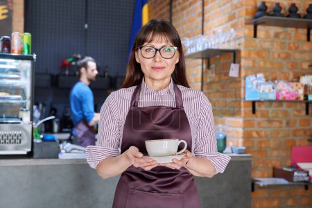 Photo for Woman in apron, food service coffee shop worker, small business owner with cup of fresh coffee, looking at camera near bar counter with male barista. Staff, occupation, entrepreneur, work concept - Royalty Free Image
