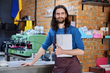 Photo for Young man in apron, food service worker, small business owner entrepreneur holding laptop looking at camera near counter of coffee shop cafe cafeteria. Staff, occupation, successful business, work - Royalty Free Image