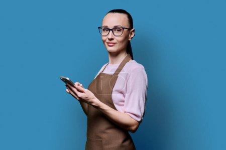 Photo for Portrait of 30s woman in apron with smartphone looking at camera on blue background. Smiling female using mobile phone texting receiving sending order. Technologies applications service small business - Royalty Free Image