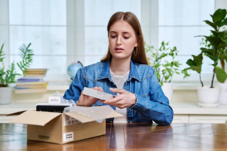 Photo for Satisfied young teen female shopper consumer sits at home and unpacks cardboard box with online purchases. Teenager girl unpacking box with new smartphone, headphones. Delivery by mail, online store - Royalty Free Image