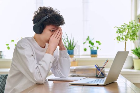 Photo for Sad upset young guy student in headphones sitting at home at desk with computer laptop, touching head with hands. Problems headaches troubles in college difficulties in study mental stress depression - Royalty Free Image
