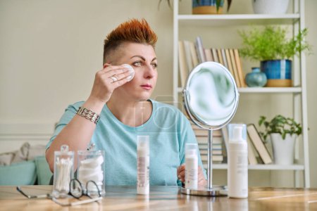 Photo for Middle-aged woman with mirror sitting at table at home. Facial anti-aging skin care products for cleansing moisturizing nourishing. Beauty age health hygiene, medicinal cosmetics for mature 40s skin - Royalty Free Image