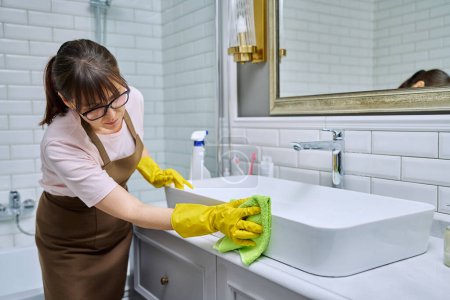 Photo for Woman in apron with detergent washcloth cleaning in bathroom, washing sink washbasin. Housewife cleaning house, cleaner service worker at workplace. Home hygiene housecleaning housekeeping housework - Royalty Free Image