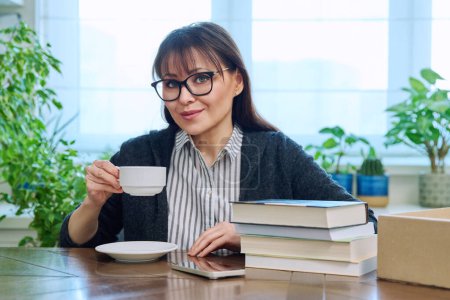 Foto de Mujer de mediana edad sosteniendo una taza de café, sentada en la mesa con un montón de libros en casa, relajada sonriendo mirando a la cámara. Ocio, estilo de vida, afición literaria, concepto de personas maduras - Imagen libre de derechos