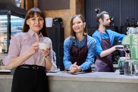 Photo for Woman customer of coffee shop near counter with cup of coffee looking at camera, restaurant workers owners at workplace. Colleagues partners in food service work, entrepreneurship, small business - Royalty Free Image