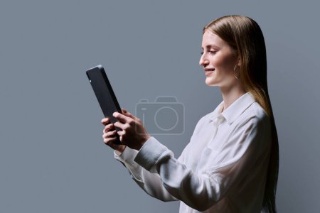 Photo for Teenage female student using digital tablet on gray background, profile view. Teenager girl 16,17, 18 years old reading looking at screen. Education Internet technology e-learning educational services - Royalty Free Image