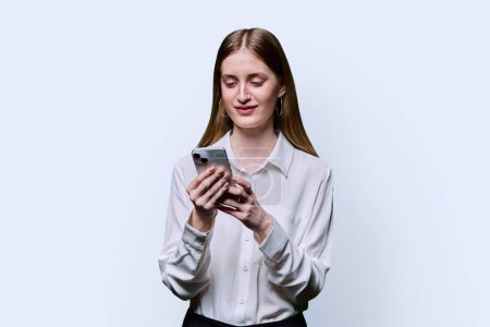 Photo for Teenage student girl 16, 17, 18 years old in white shirt holding smartphone in hands, texting looking at screen on white studio background. Education technology high school college lifestyle youth - Royalty Free Image