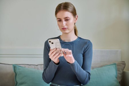 Serious calm female teenager using smartphone, sitting on couch at home, girl 16,17, 18 years old texting reading messages. Modern digital technologies for communication, leisure, learning, shopping