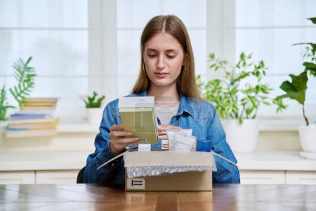 Zufriedene junge Kundin sitzt zu Hause und packt Karton mit Online-Einkäufen aus. Teenagermädchen beim Auspacken von Schachteln mit Kosmetikartikeln. Lieferung per Post, Online-Shop