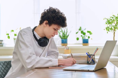 Téléchargez les photos : Jeune étudiant masculin assis au bureau à la maison à l'aide d'un ordinateur portable. Homme 19-20 ans étudiant à la maison, utilisant un ordinateur pour l'éducation loisirs communication - en image libre de droit