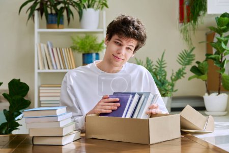 Téléchargez les photos : Satisfait jeune acheteur de client masculin assis à la maison déballer colis boîte en carton avec de nouveaux livres, achats en ligne. Livraison par courrier, librairie internet - en image libre de droit
