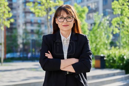 Photo for Portrait of middle-aged business confident woman in suit, with crossed arms, posing outdoors in urban style. Office employee worker businesswoman teacher entrepreneur economist banker financier lawyer - Royalty Free Image