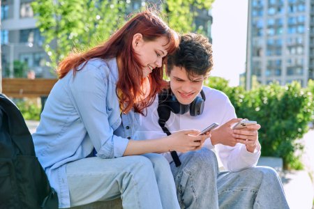 Foto de Adolescentes amigos jóvenes chico y chica universitarios estudiantes universitarios sentados al aire libre en los escalones del campus hablando riendo con un teléfono inteligente. Tecnología, concepto de estilo de vida - Imagen libre de derechos